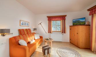 A cozy living room with an orange couch and a small table. In the background, a TV and a wooden cabinet can be seen.