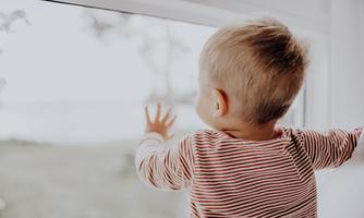 A small child stands by the window and looks outside. It presses its hand against the glass and gazes curiously at the landscape.