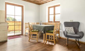 A cozy dining area with a table and chairs, surrounded by bright windows. The room features a warm wooden ceiling and a modern armchair.