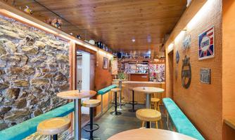 A cozy interior with a wooden ceiling and stone wall. There are round tables and a bar in the background.