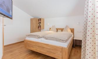 A cozy bedroom with a double bed and wooden furniture. A television hangs on the wall, and there is a closet for storage.