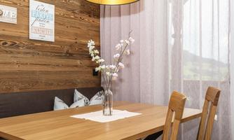 A cozy dining area with a wooden table and a vase full of flowers. The windows are decorated with light curtains, and there is a warm ceiling lamp.