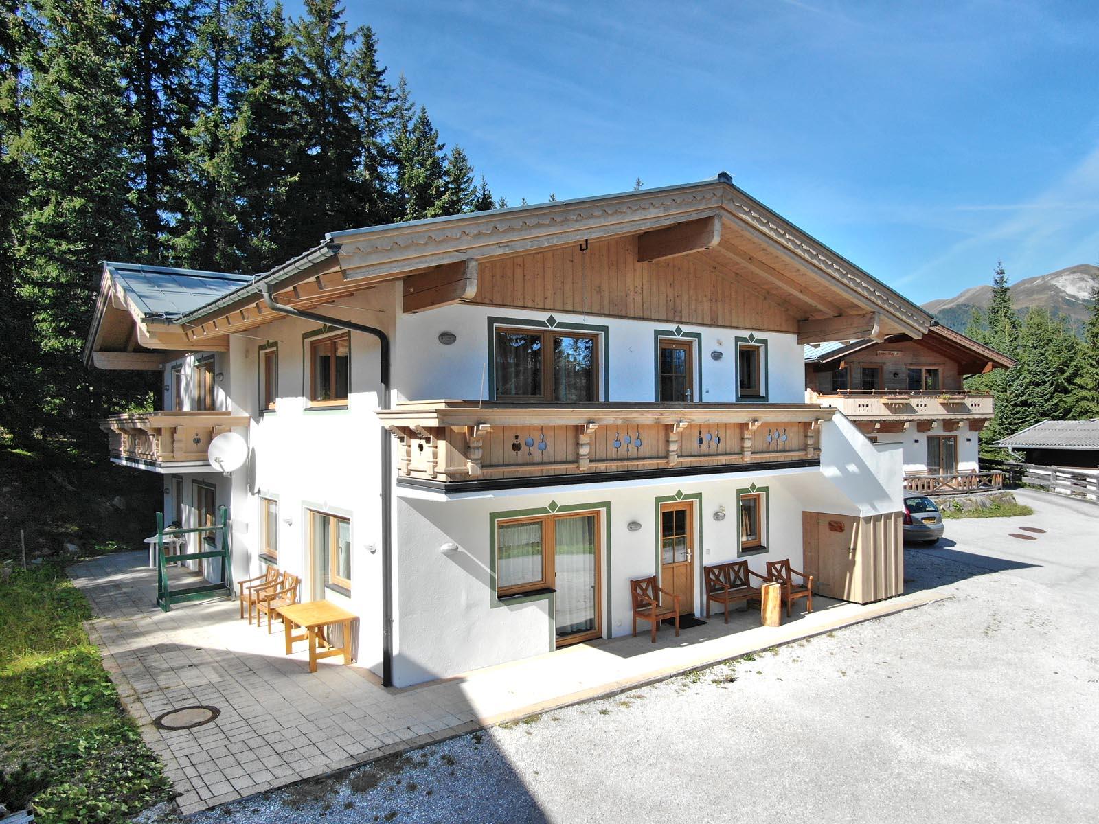 A modern alpine house with wooden cladding and an adjacent balcony. Surrounded by green trees and a clear sky.