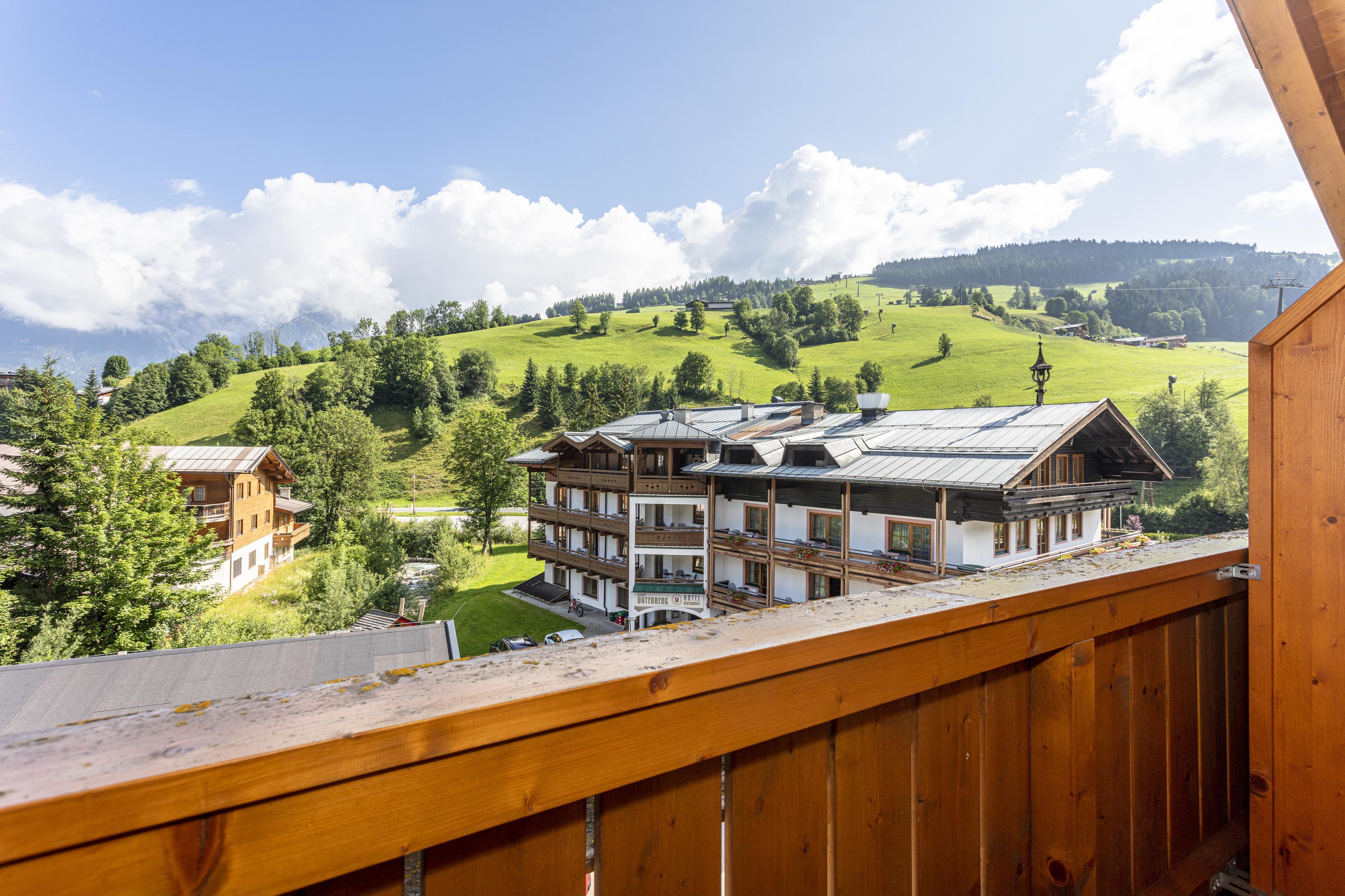 A picturesque view of green hills and a modern hotel in the Alps. The scene is surrounded by blue sky and white clouds.