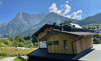 A modern wooden house is picturesque in the mountains. In the background, there are high mountains and a blue sky.