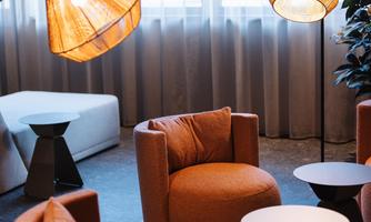 A cozy lounge area with orange armchairs and modern lamps. In the background, bright curtains and a gray carpet are visible.