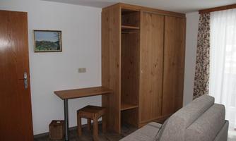 A simple room with a desk, a chair, and a wooden wardrobe. A painting hangs on the wall and the light streams through the window.