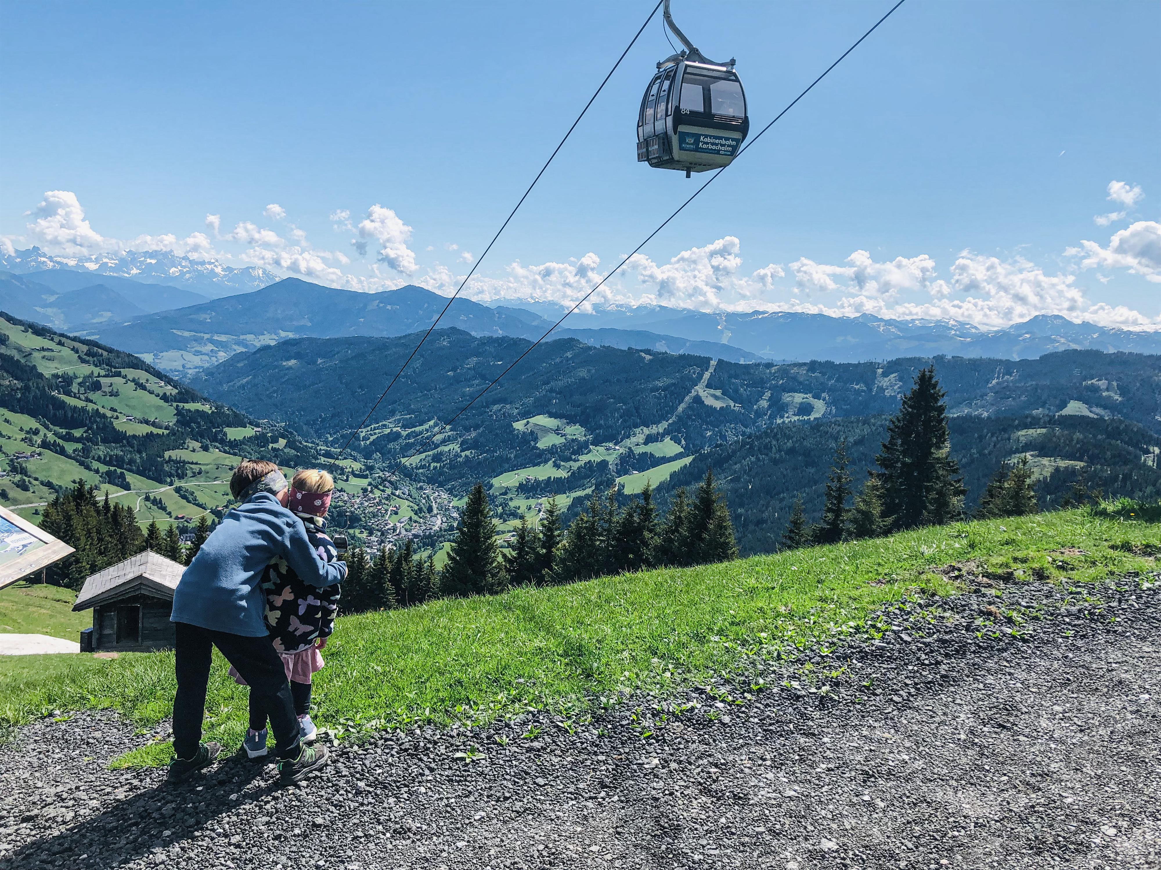 In the picture, two children are standing on a meadow and looking at the mountains. In the background, there is a cable car as well as a beautiful, green landscape.