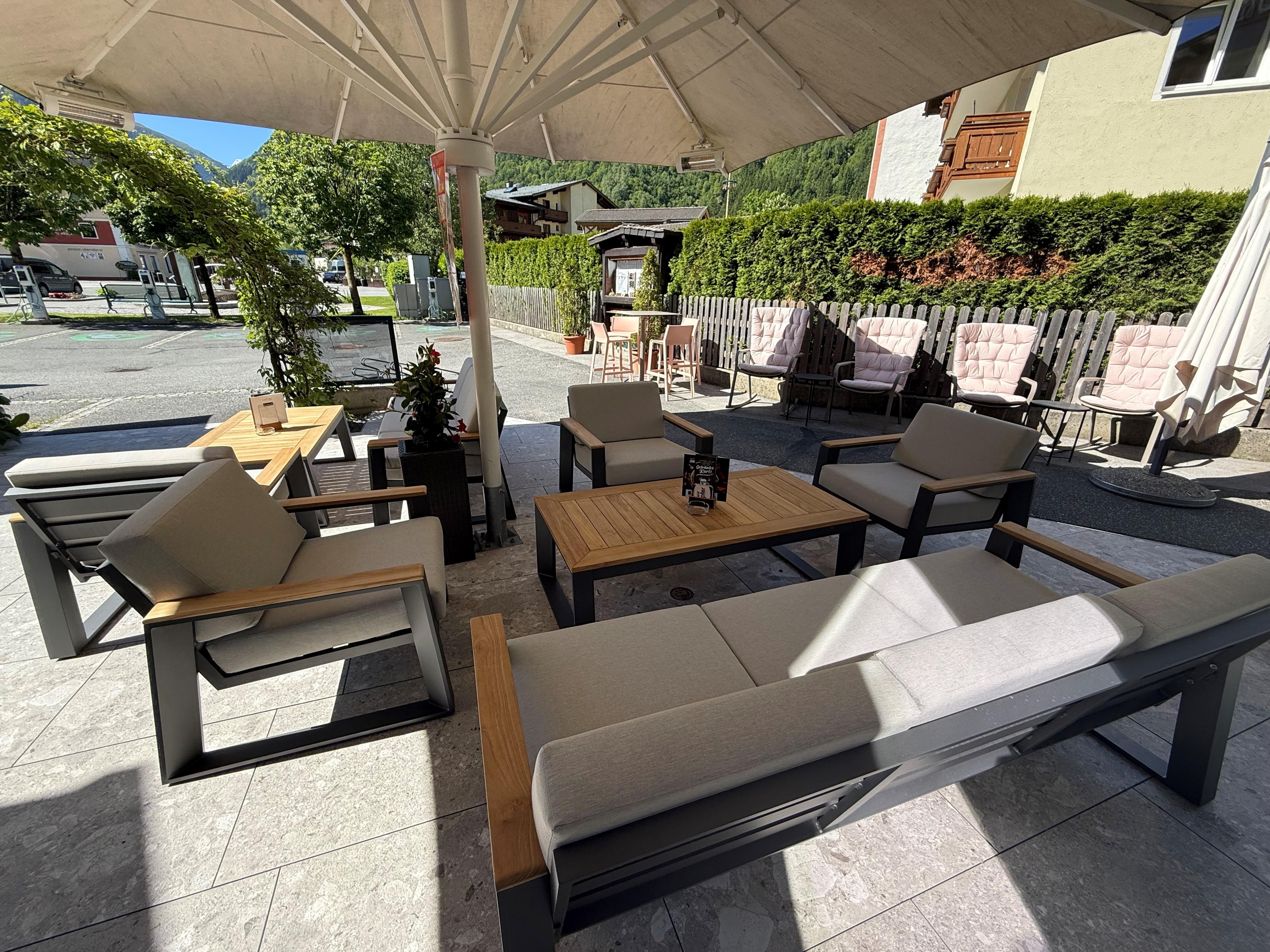 A cozy outdoor area with modern furniture. There is a large sun umbrella and a table with chairs, surrounded by green plants.