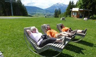 Three people are relaxing on lounge chairs in a beautiful mountain landscape. In the background, the mountains and a small building are visible.