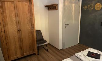 A simple bedroom with two beds, a wooden wardrobe, and a chair. The walls are painted in neutral tones.