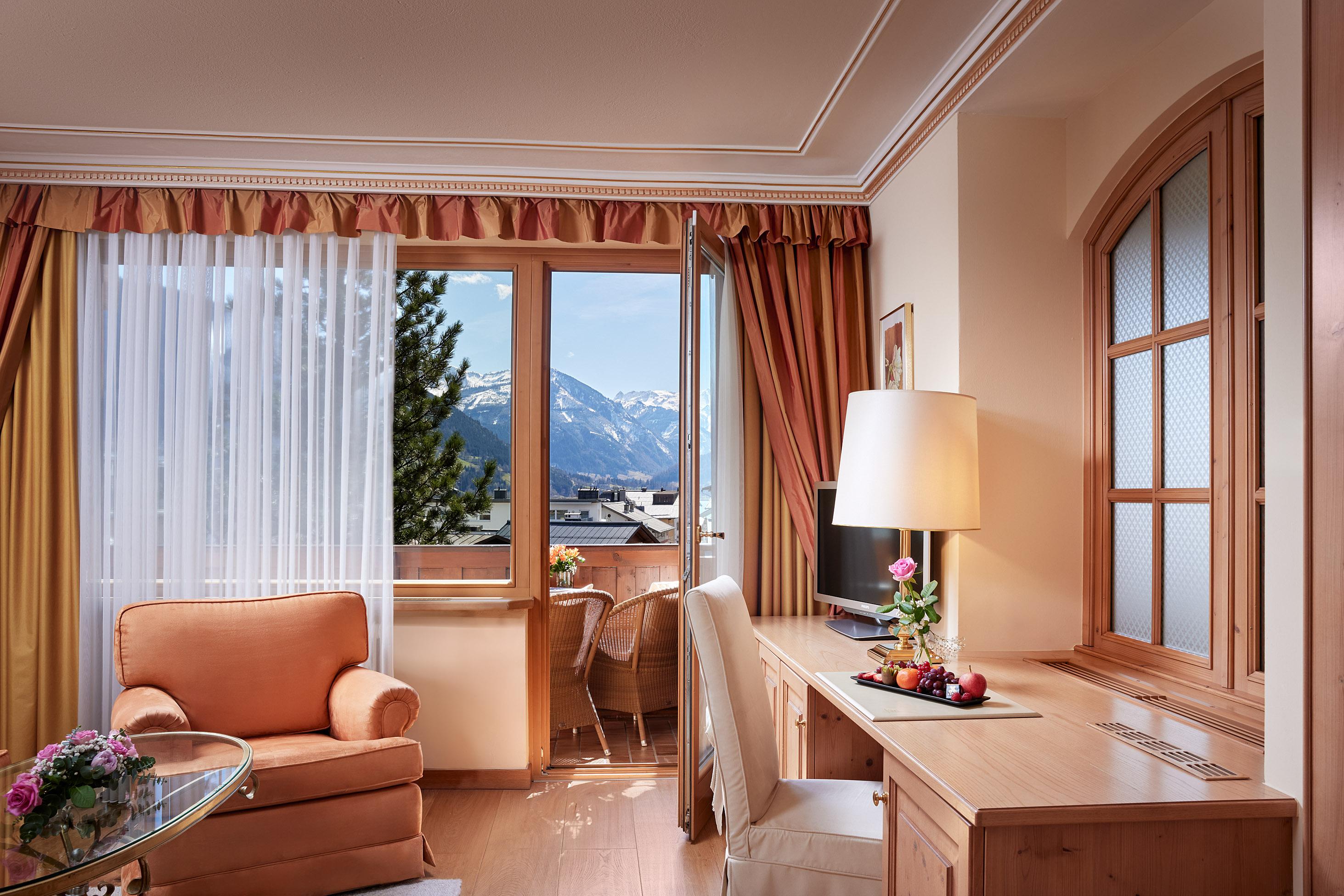 A stylish hotel room with a comfortable armchair and a desk. The large windows offer a beautiful view of the mountains.