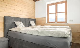 A modern bedroom with a large bed and two pillows. The walls are made of light wood, and a window with a view provides daylight.