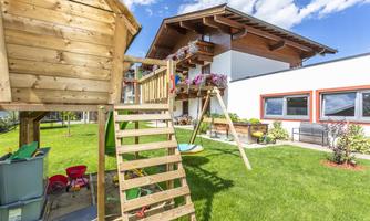 A playground with a wooden tower and a slide, surrounded by green grass. In the background, a bright house with flower boxes can be seen.
