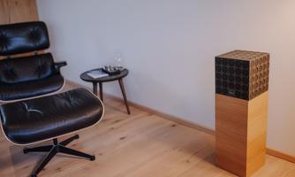 A stylish room with a comfortable black armchair and a modern speaker on a wooden stand. The floor is made of light wood, and there is a small table next to it.