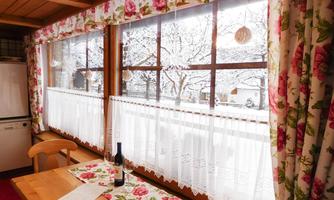 A cozy room with a large window overlooking a snowy landscape. The windows are adorned with blooming curtains and a table stands in the foreground.