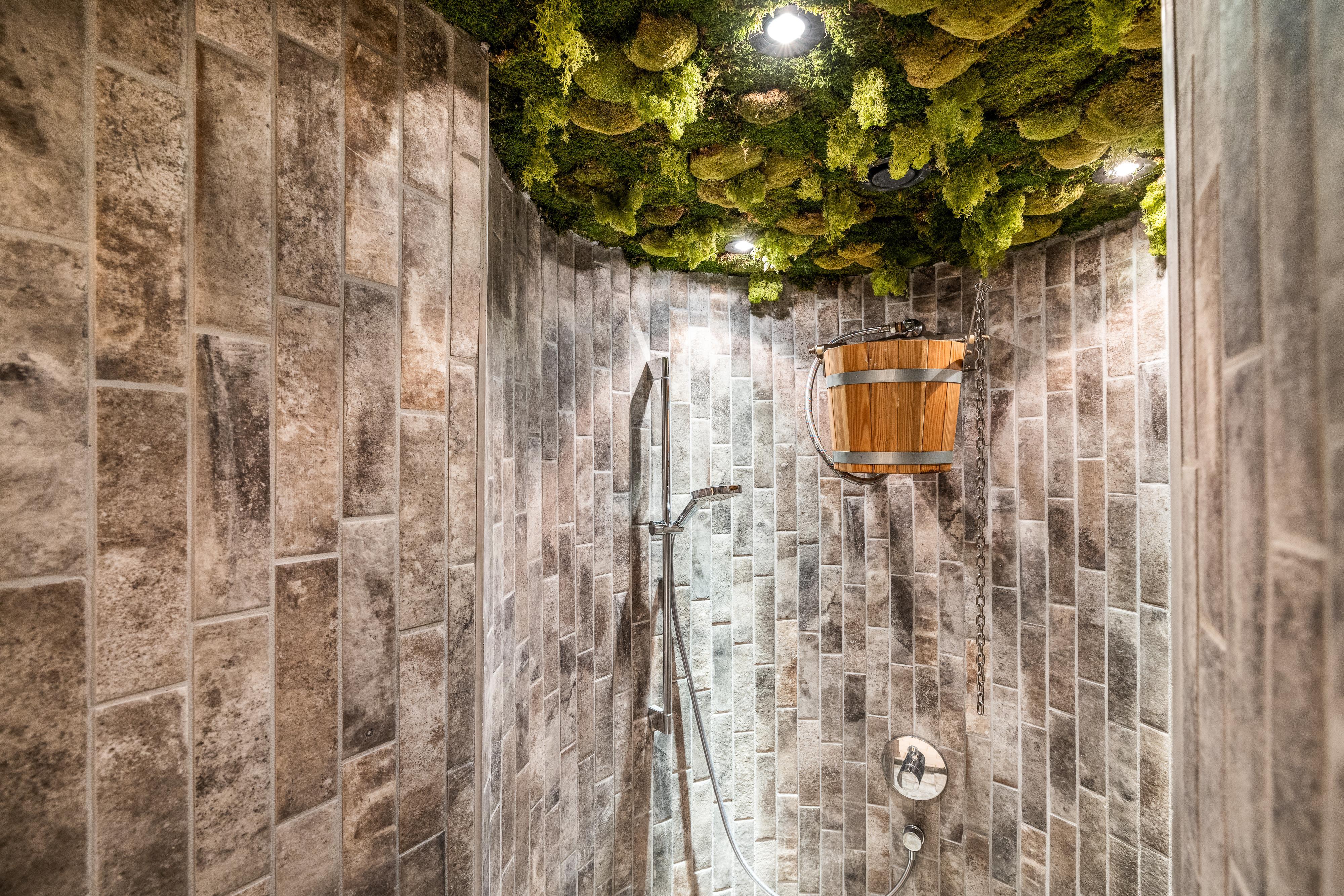 A modern shower with gray tiles and a green ceiling made of moss. Above the shower hangs a wooden water tank.