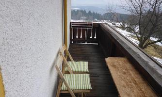 A balcony with two chairs and a wooden table. In the background, a wooded landscape and a cloudy sky are visible.