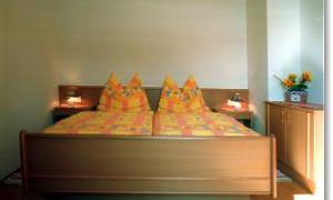 A cozy bedroom with a double bed and colorful pillows. Next to the bed are bedside tables and a dresser, decorated with a vase of flowers.