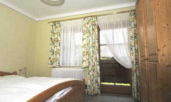A cozy bedroom with a wooden bed and floral curtains. The window offers a view of a balcony.