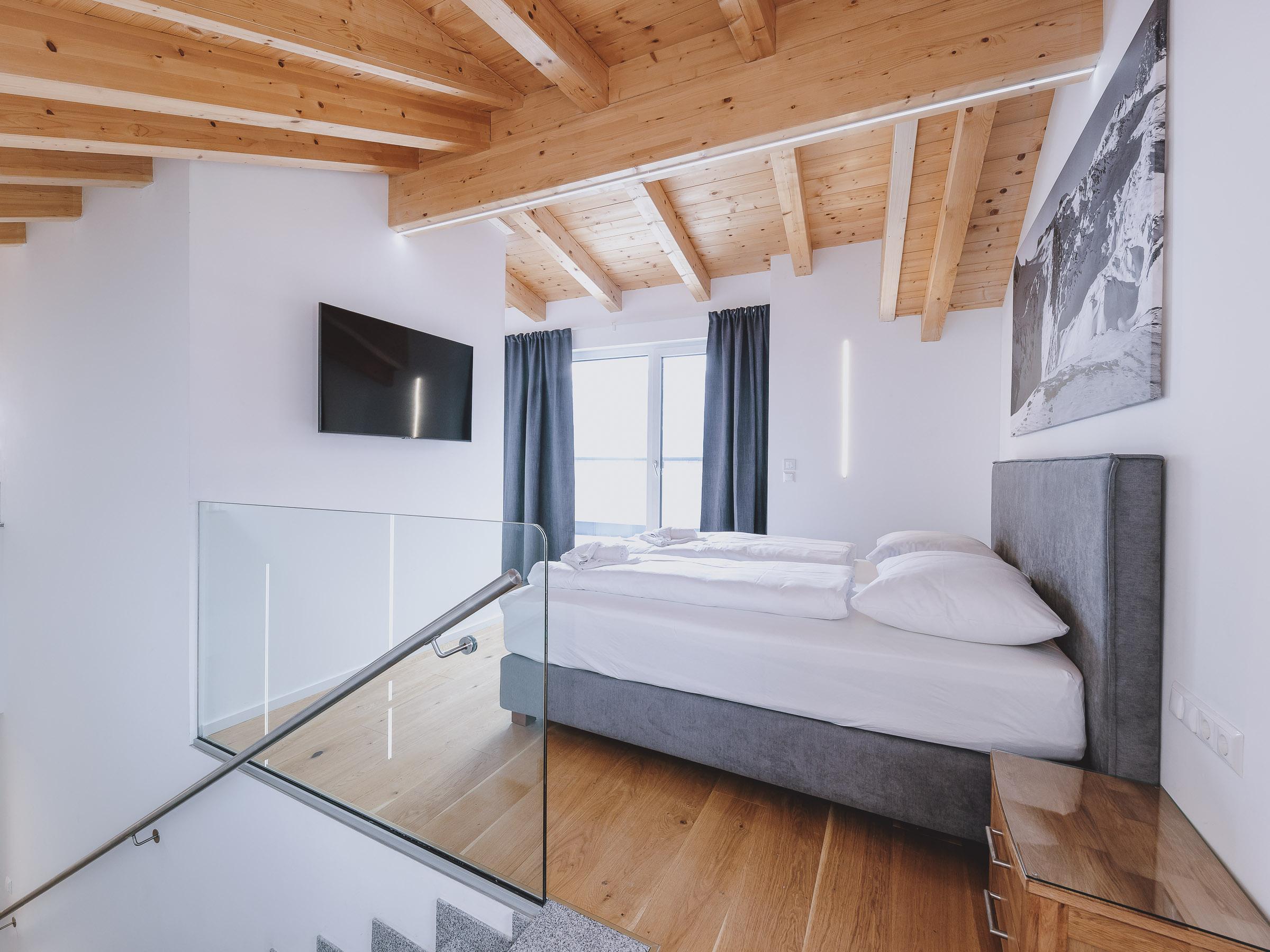 A modern bedroom with wooden ceilings and large windows. The room is bright and stylishly decorated, featuring a large bed and a TV.