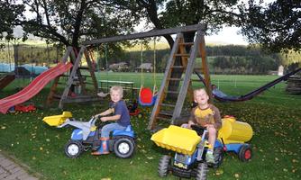 Two children are happily playing on a playground with slides and swings. They are riding small tractors and enjoying their time outdoors.