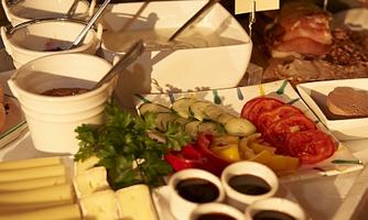 A rich buffet with various types of cheese, fresh vegetables, and cold cuts. It also includes sweet spreads and rolls.
