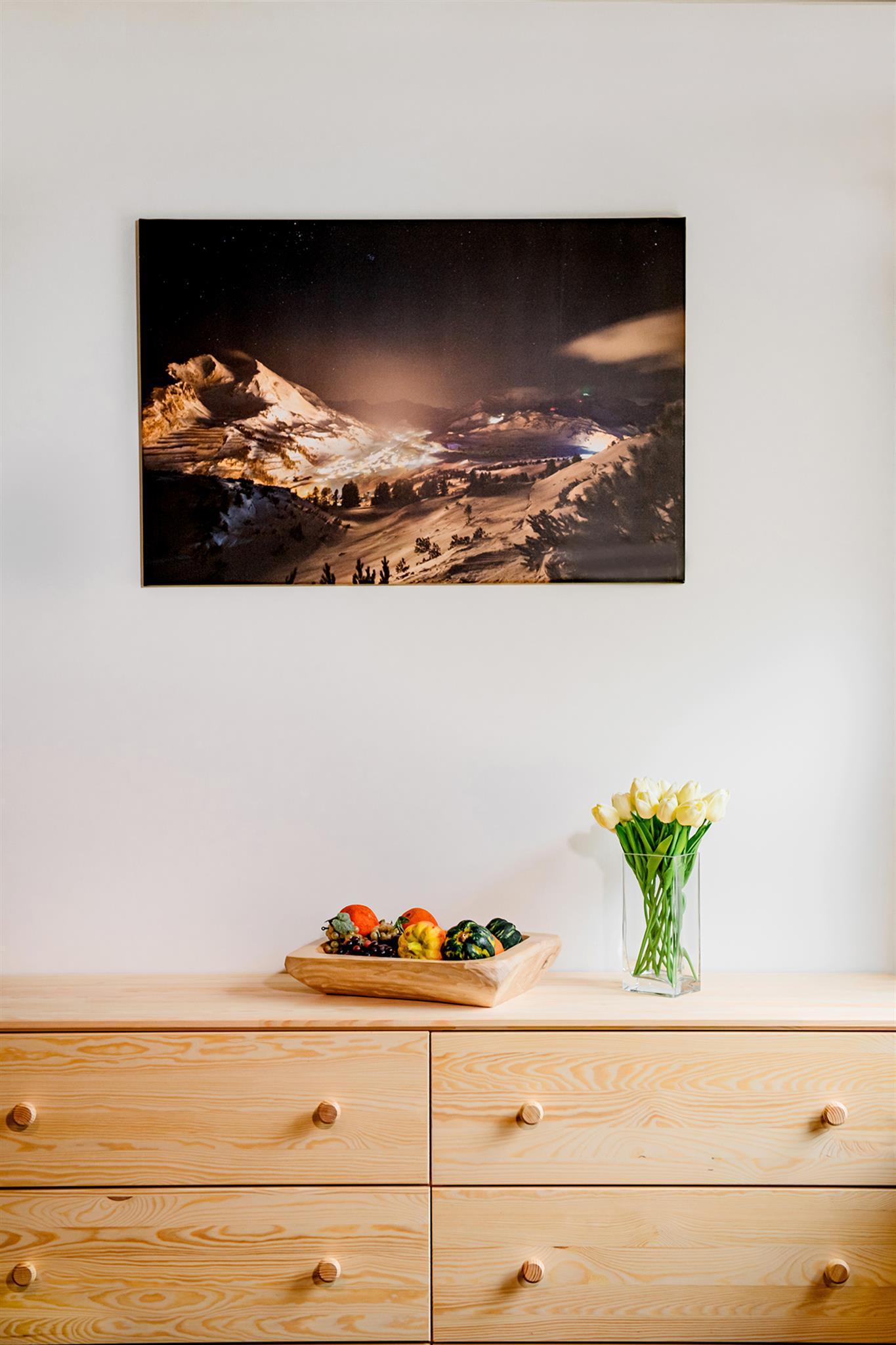 A modern room with a wooden chest of drawers. On the chest of drawers sits a bowl of fruit and a bouquet of flowers in a vase. A landscape painting hangs on the wall.