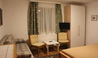A simple room with two chairs, a small table, and a sofa. There is a television and a wardrobe, along with curtains and a rug.