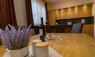 A modern dining area with a wooden table and stylish decor. In the background, a kitchen and windows with curtains are visible.