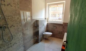 A modern bathroom with a glass shower and a window. The walls are in neutral colors, and a green door adds a splash of color.