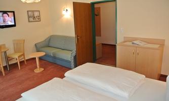 A cozy hotel room with a double bed, a sofa, and a small table. A television hangs on the wall, and there is an open door to another room.