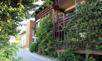 A modern building, surrounded by green trees and bushes. The street is narrow and runs along the facade.