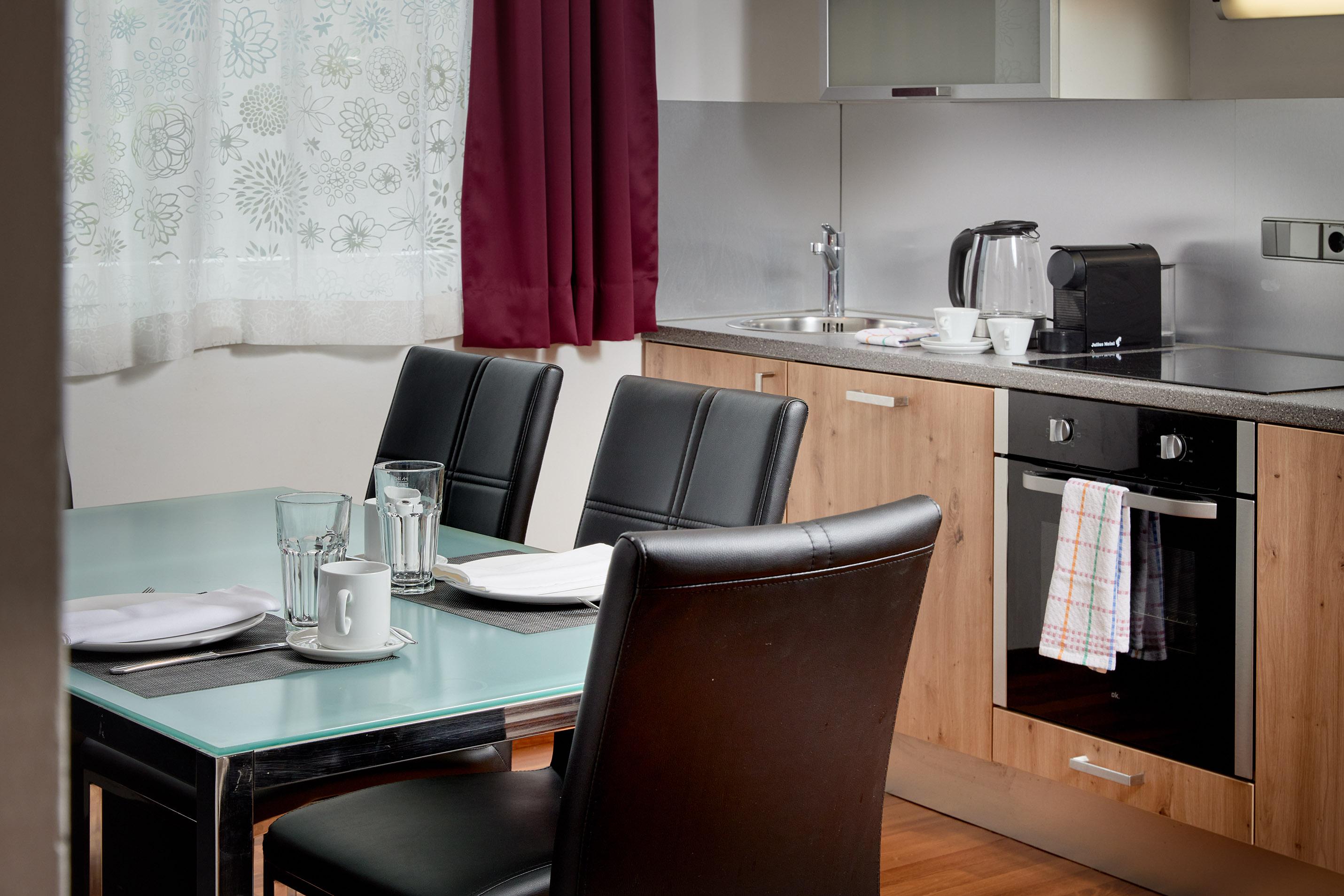 A modern kitchen with a dining table and black chairs. The room features wood and stainless steel accents as well as a bright window front.