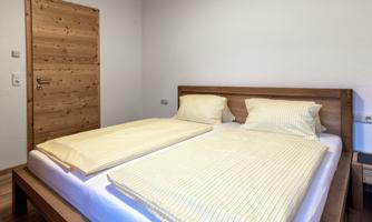 A cozy bedroom with a double bed and light yellow bedding. The walls are kept neutral and a wooden door leads outside.