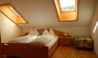 A cozy bedroom with a double bed and two windows in the roof. The floor is made of wood and there is a small chest of drawers as well as a colorful seating area.