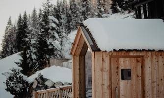 A wooden house stands in the snow, surrounded by snow-covered trees. The cold atmosphere and the natural surroundings give the place a cozy ambiance.