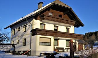 A charming three-story house with wooden elements in the winter snow. The surroundings are quiet, with trees in the background and a clear blue sky.