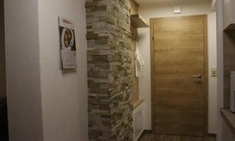 A modern hallway with a stone wall and a wooden front door. A calendar is hanging on the wall, and the floor is covered with wooden planks.