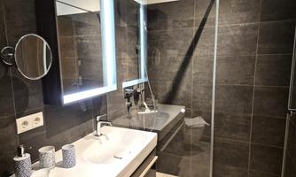 A modern bathroom with dark tiles and a glass shower. The sink has a lighted mirror and elegant accessories.