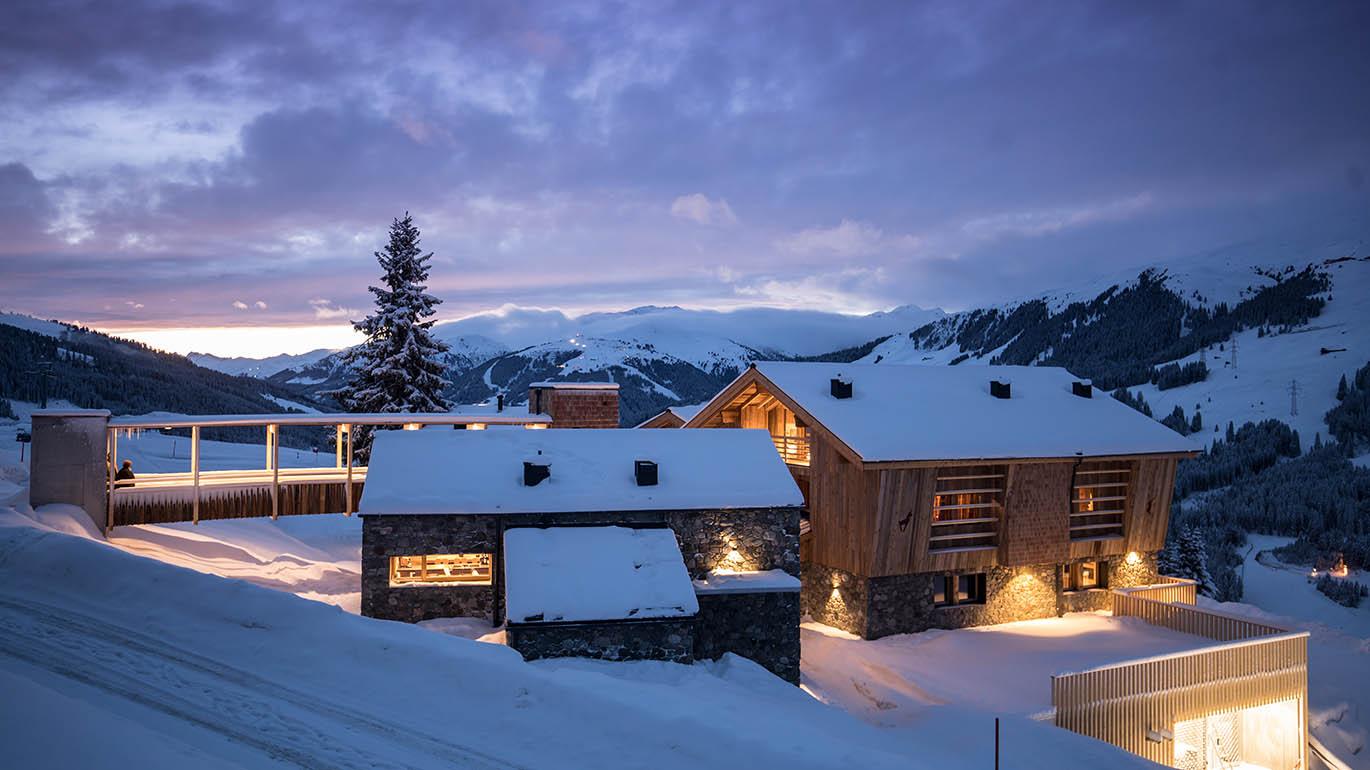 A modern chalet in the snow with mountains in the background. The sky is tinted in soft colors of dusk.