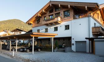 A modern house with traditional wooden design in a mountainous setting. There are covered parking spaces and a well-maintained garden landscape.