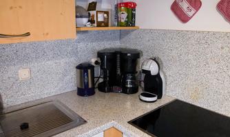 A modern kitchen corner with a kettle, a coffee machine, and an induction cooktop. The countertop is made of gray stone and there are some kitchen utensils in the background.