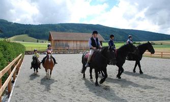 Riders and children on horses in a riding arena. In the background, green meadows and mountains can be seen.