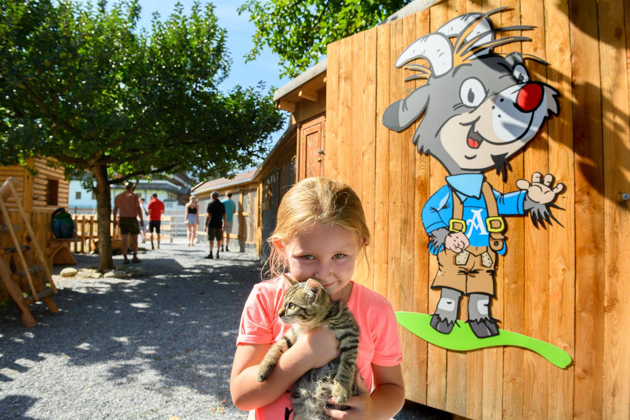 A girl is holding a kitten in her arms. In the background, there is a wooden building and several people.