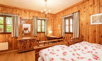 A cozy bedroom with wood-paneled walls and a large window. The bed is covered with a floral duvet and a wooden wardrobe stands next to a vanity table.