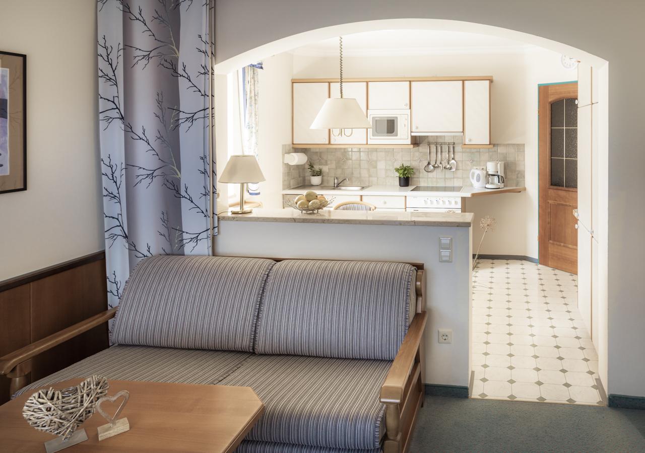 A cozy living room with a sofa and a table. The open kitchen in the background is bright and modernly furnished.