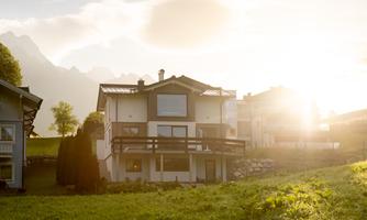 A modern house in a green setting with mountains in the background. The sun is shining bright, creating a warm atmosphere.