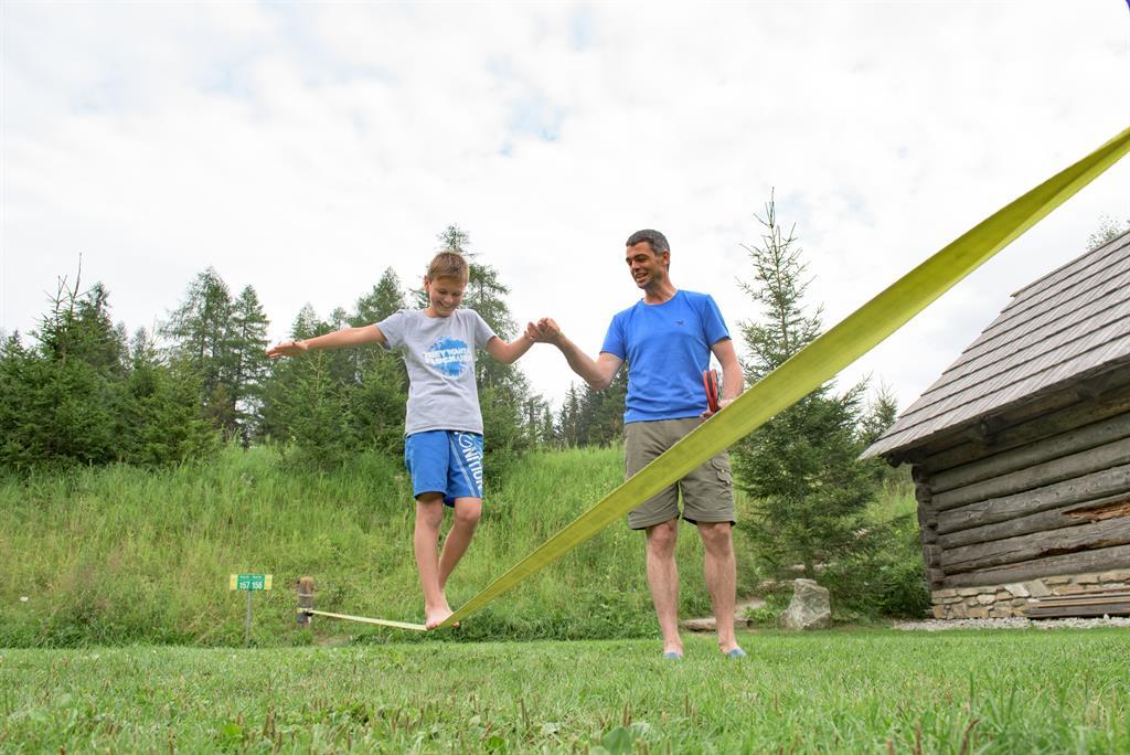 A boy is balancing on a slackline while an adult stands beside him. In the background, trees and a wooden house are visible.