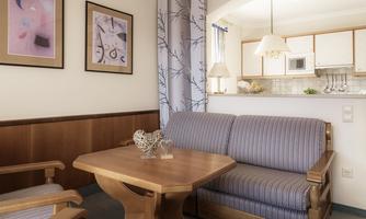 A cozy living room with a couch and a wooden table. The room has modern artworks on the wall and an open kitchen in the background.